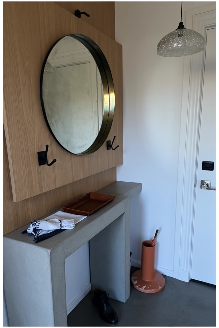 Wooden wall panel in entryway with round mirror, hooks, slim concrete console table, and umbrella stand