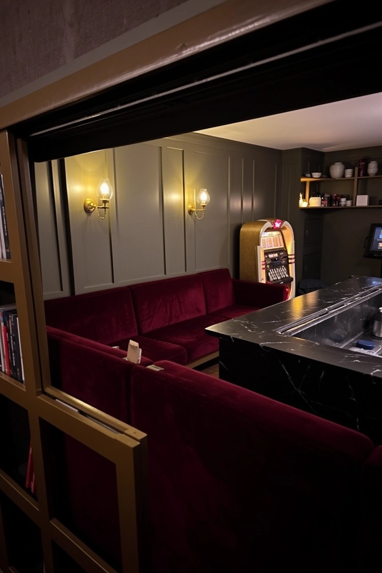 Red velvet L-shaped booth seating in a home pub corner, framed by golden bookshelves, with black marble bar top, retro jukebox, and warm wall lamps