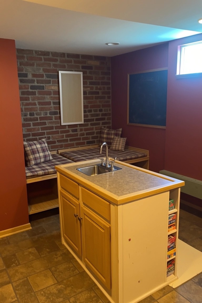 Basement pub room with red exposed brick wall behind wooden kitchenette island, sink, and built-in bench seating with plaid cushions