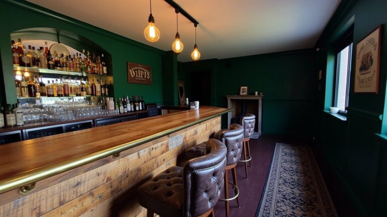 Rustic wooden home bar counter with leather stools against dark green walls, lit bottle shelves, and a fireplace in the corner