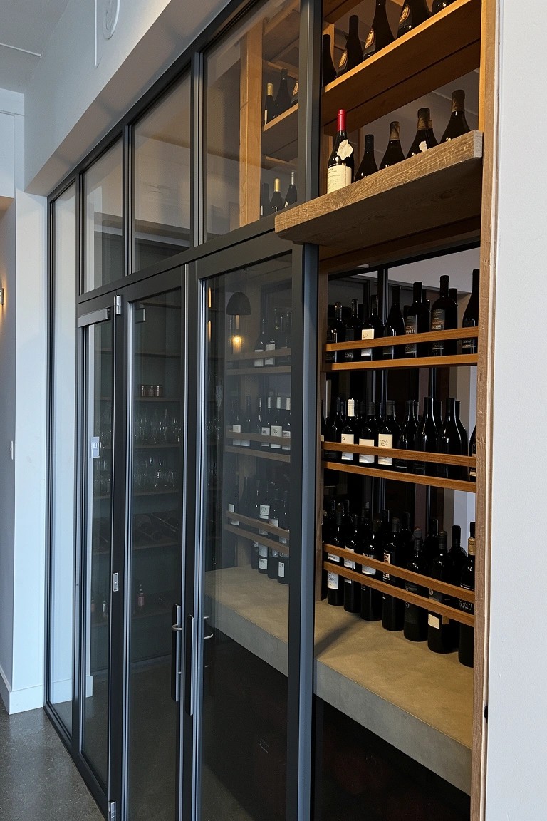 Glass-fronted wine storage room with wooden shelves full of bottles, black sliding doors, and concrete flooring in a modern home.