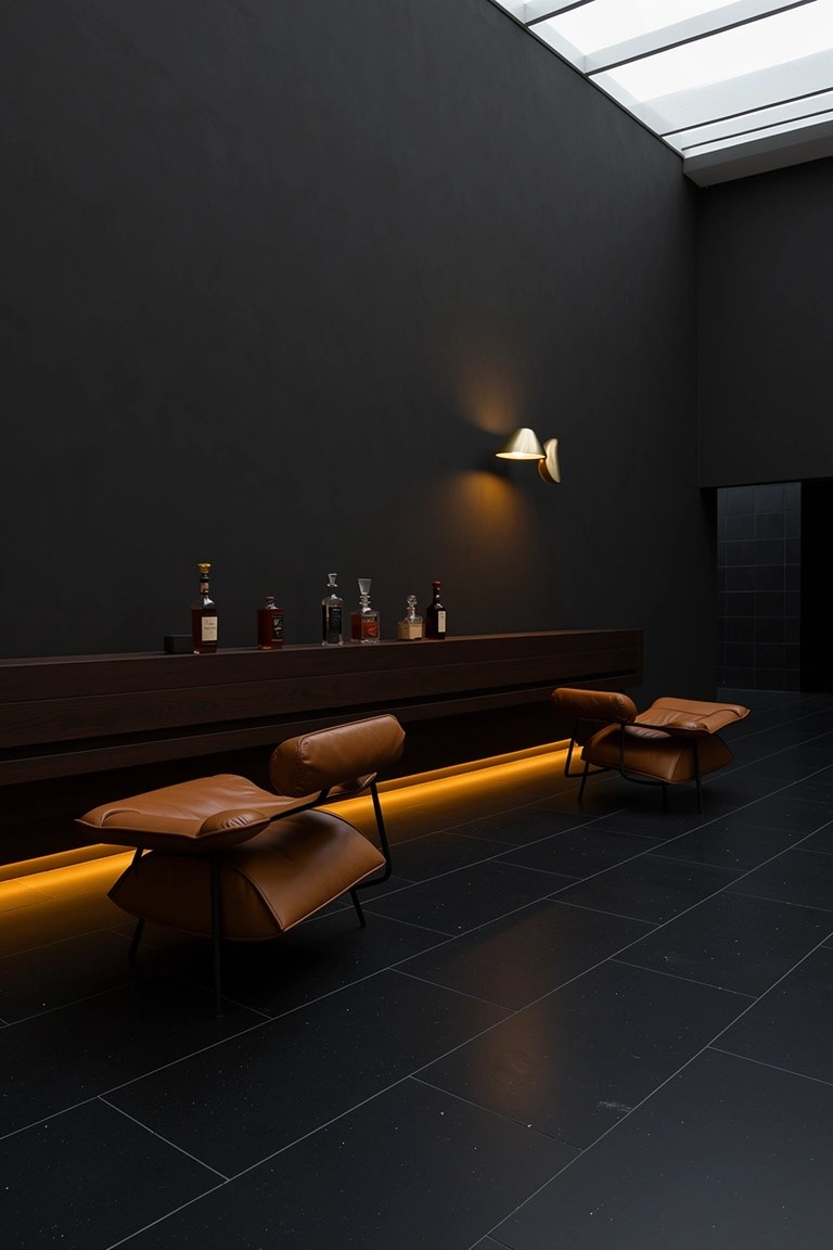 Tan leather lounge chairs with warm underlighting next to a wooden bar shelf holding bourbon bottles in a dark-walled room