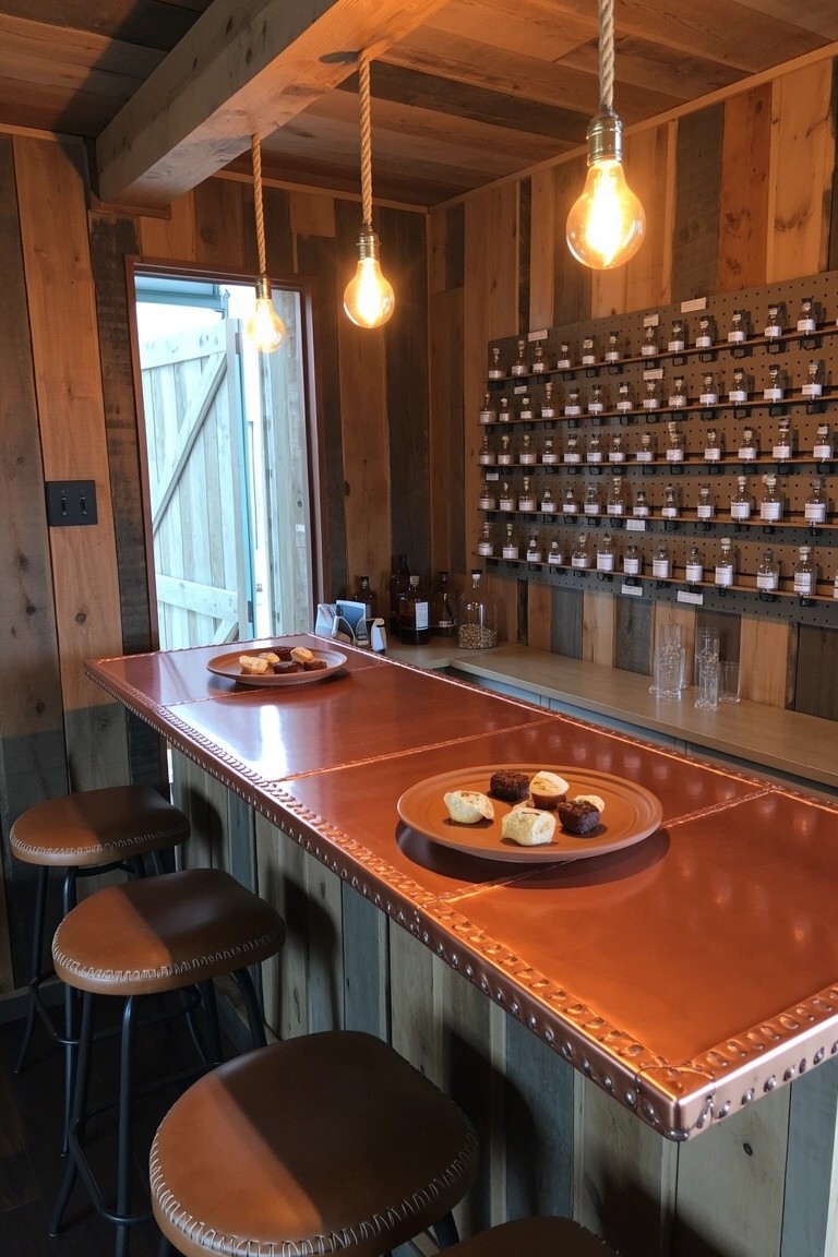 Rustic bar in wooden room with wall shelves of labeled glass bottles above copper countertop and leather stools