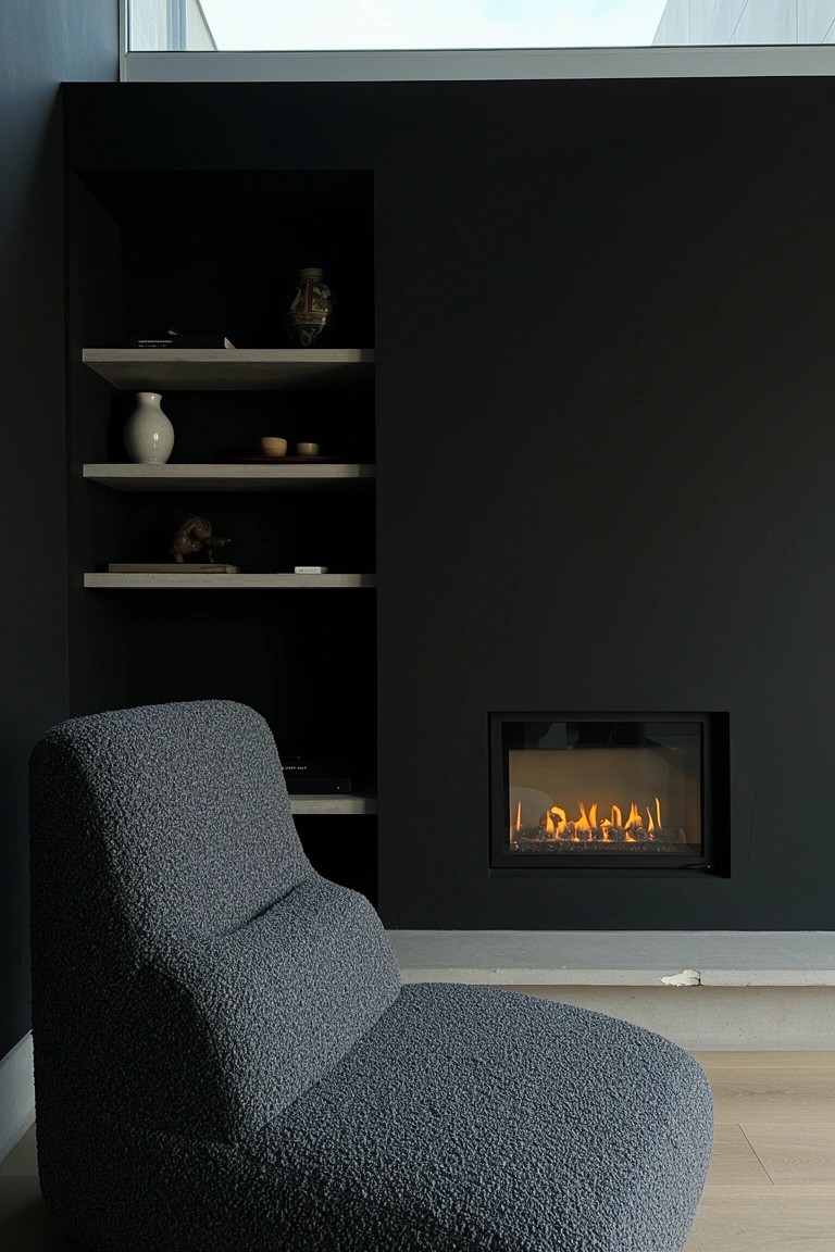 Dark feature wall with slim built-in fireplace flanked by floating shelves holding simple decor and a plush textured gray armchair in front
