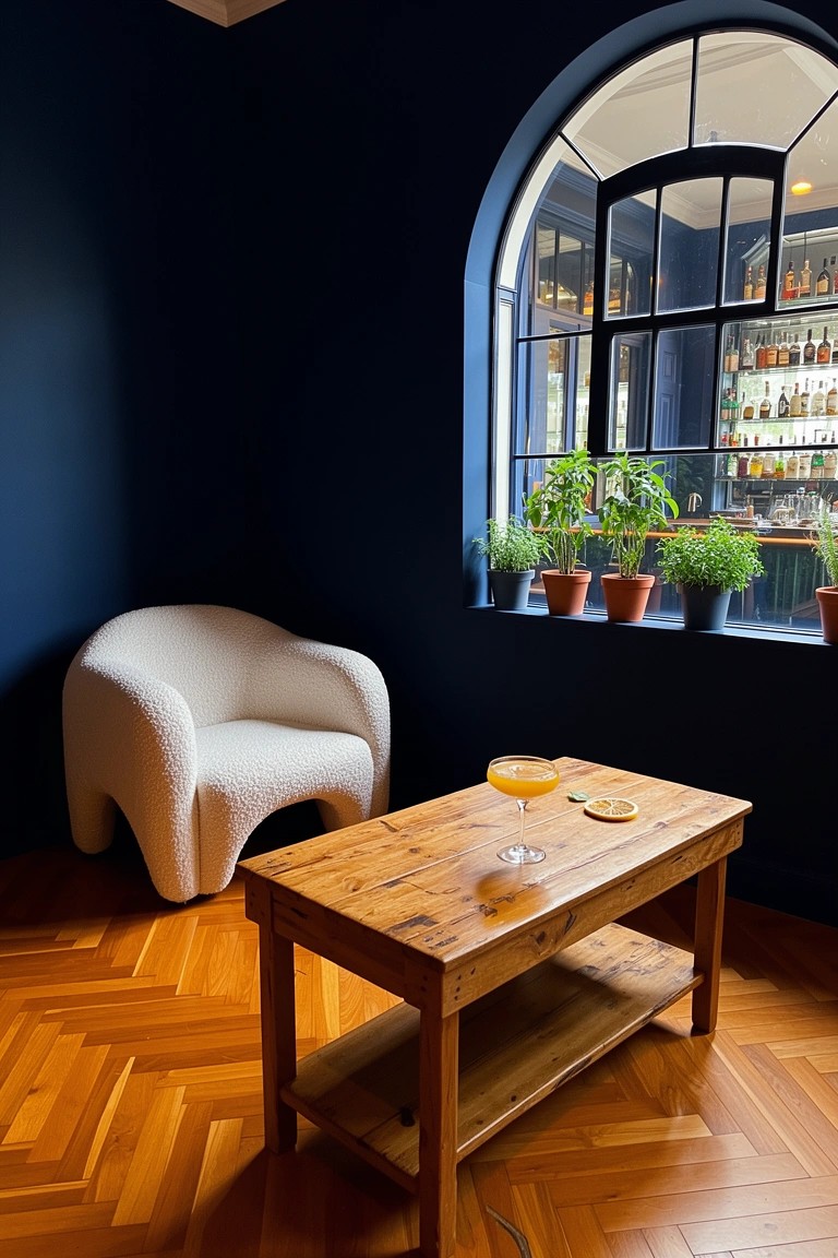 Navy blue corner nook with white plush armchair, rustic wood coffee table holding drinks, and arched window with plants and bar view
