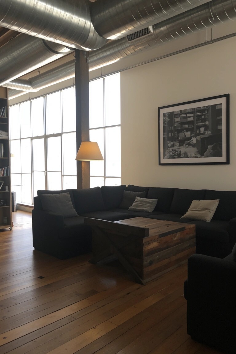 Industrial man cave lounge with exposed silver ducts overhead, black L-shaped sofa around a wooden coffee table, and large windows