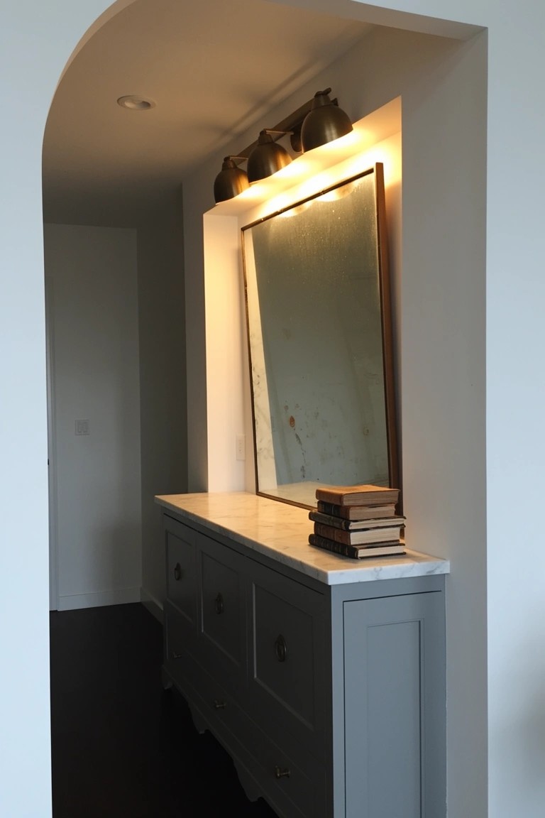 Gray console table with white marble top, stacked books, and large mirror in arched white hallway niche lit by brass sconces