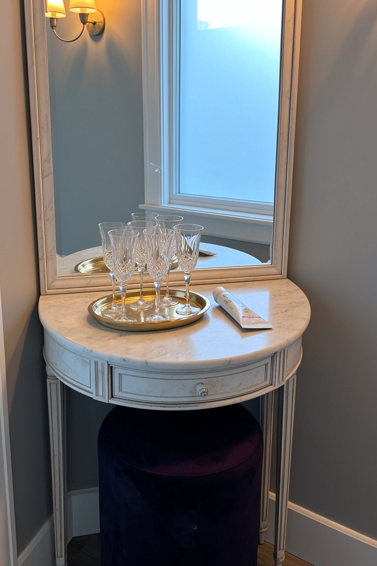 Curved white marble console table with gold tray of champagne flutes and folded napkin, purple velvet stool underneath, next to a mirror in a soft gray room