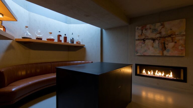 Modern whiskey room corner with wooden wall shelves displaying assorted bottles above a black bar table with a decanter, leather sofa nearby, and linear gas fireplace on neutral walls