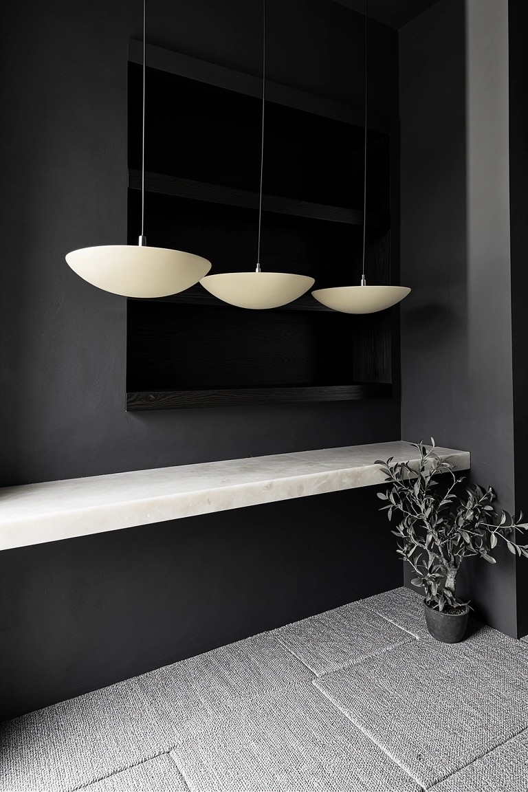 Dark modern whiskey room nook with floating white marble shelf, three cream bowl pendant lights hanging above, built-in black recess, and potted olive plant on gray floor
