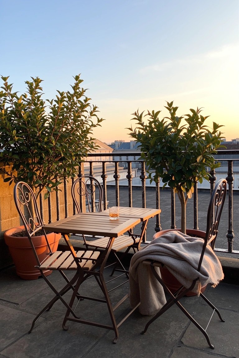 Wrought iron bistro table and two chairs on a balcony with potted plants and a draped blanket