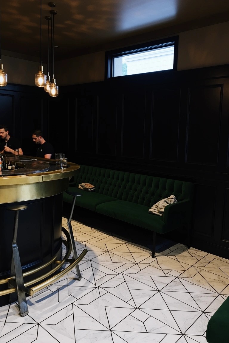 Curved brass bar counter with black stools and green velvet sofa against dark wood paneling in a man cave bar area