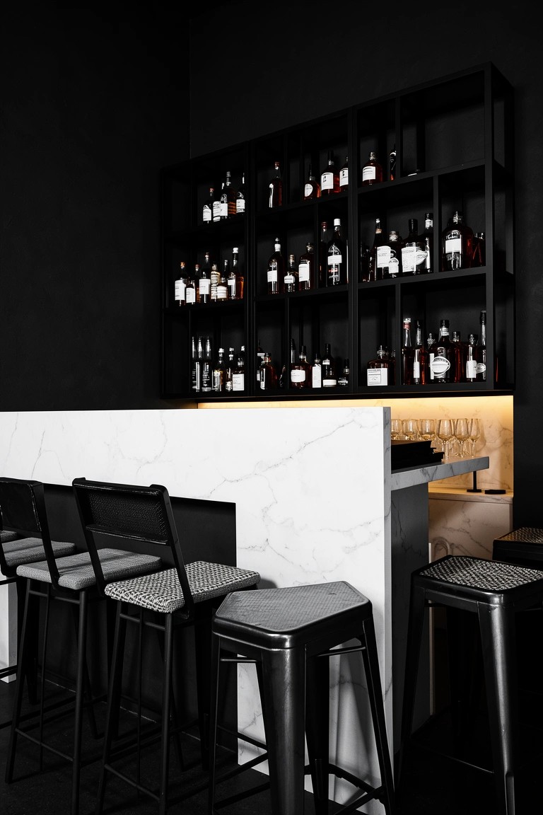Sleek black bar counter with white marble top and wall-mounted dark shelves full of whiskey bottles