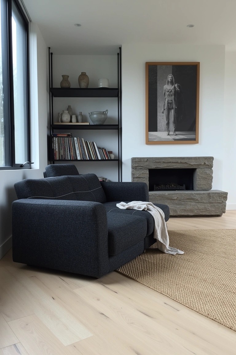 Modern man cave corner featuring a dark gray armchair with light throw beside a low stone gas fireplace, tall black metal open shelves displaying books, ceramics, and a large black-and-white portrait, seagrass rug on light wood floors