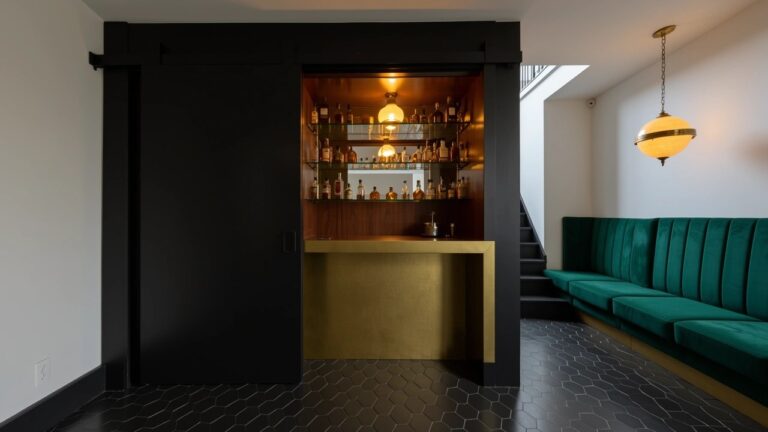 Open black sliding door revealing a cozy home speakeasy bar with bottle shelves, wood counter, and green bench