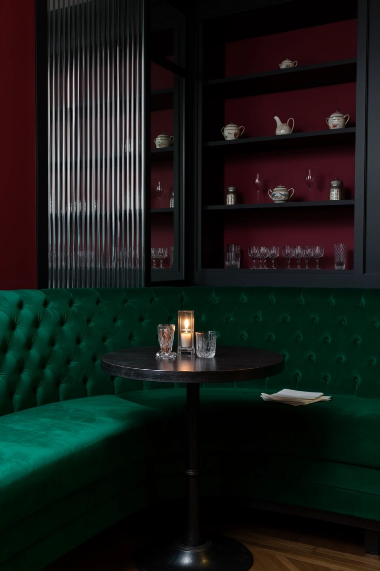 Emerald green tufted velvet L-shaped banquette around a small black metal table with candles, glasses, and napkin in a deep red room with shelved glassware