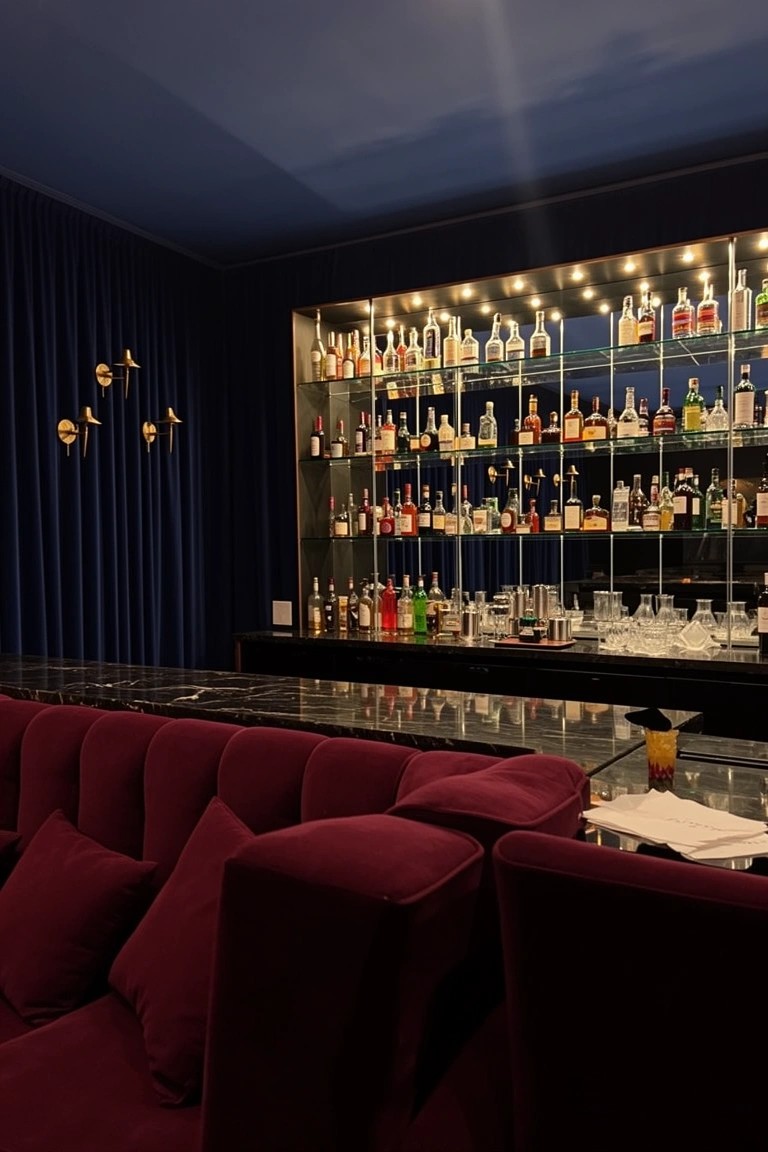 Navy-walled basement bar with tall backlit shelving full of liquor bottles, black marble counter, and maroon velvet seating