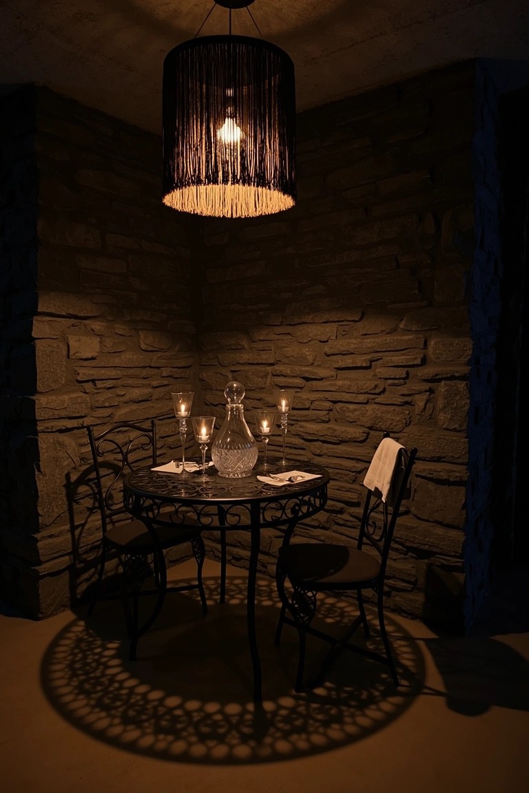 Small black wrought iron bistro table and chairs in a dark stone corner, set with candles, glass carafe, and napkins under a fringed pendant lamp