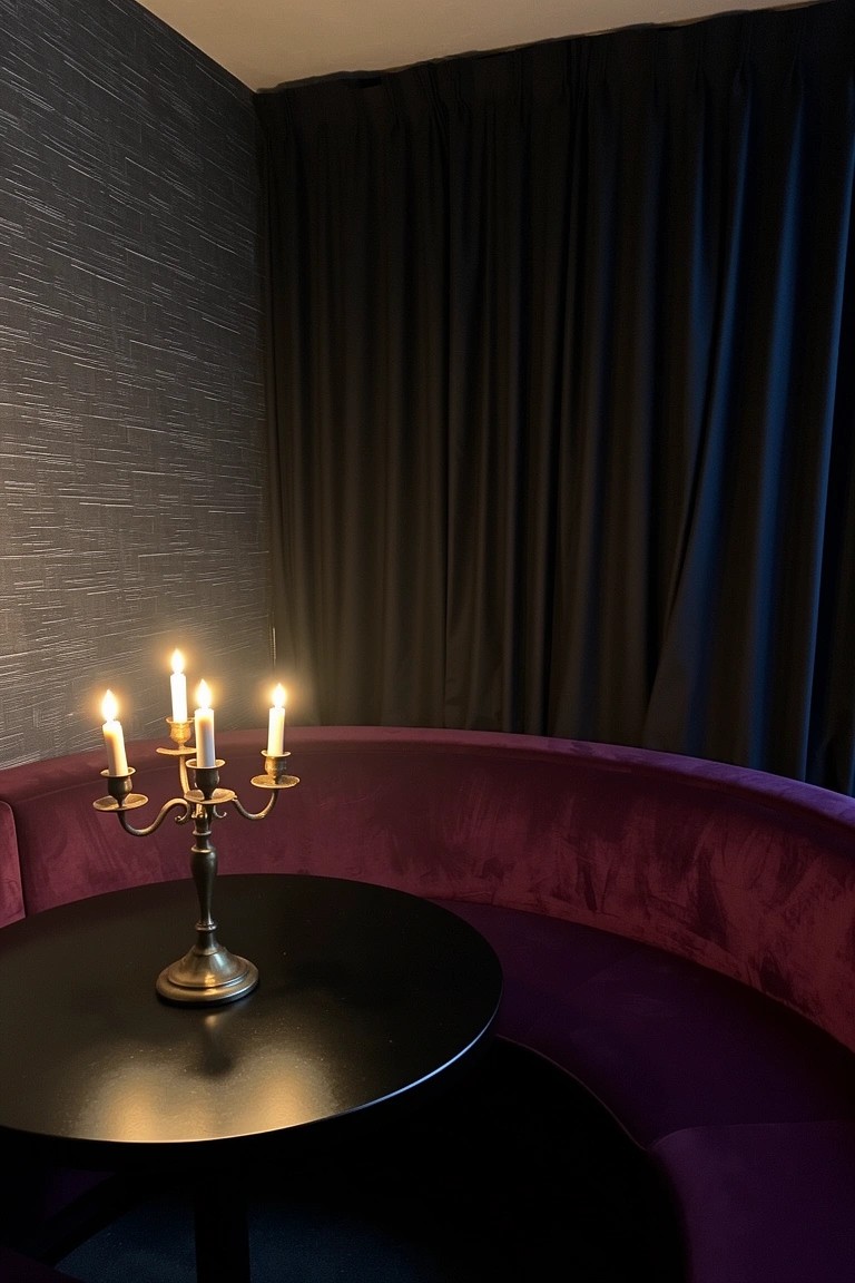 Purple velvet curved banquette beside a small black round table holding a brass candelabra with lit candles in a dark moody room corner