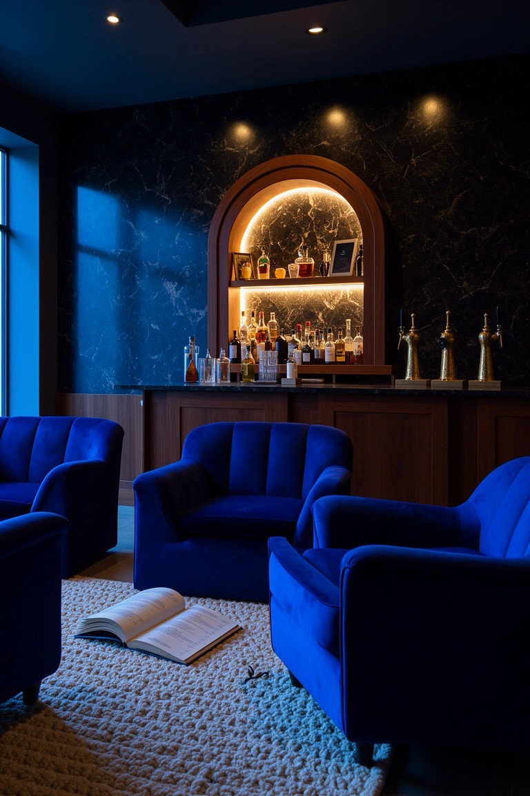 Dark home bar area with wooden cabinet featuring arched lit niche displaying whiskey bottles and glassware, surrounded by blue velvet armchairs on a rug