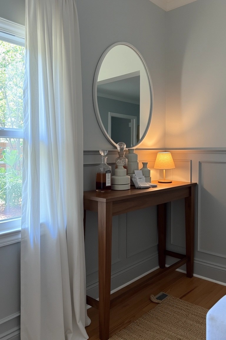 Wood console table in gray corner with oval mirror above, small lamp, stacked white ceramics, bottles, and sheer window curtains