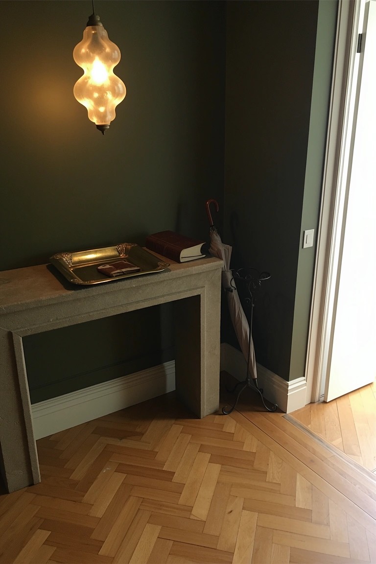 Concrete console table in green entryway corner with herringbone parquet floor, silver tray, book, and umbrellas by open door