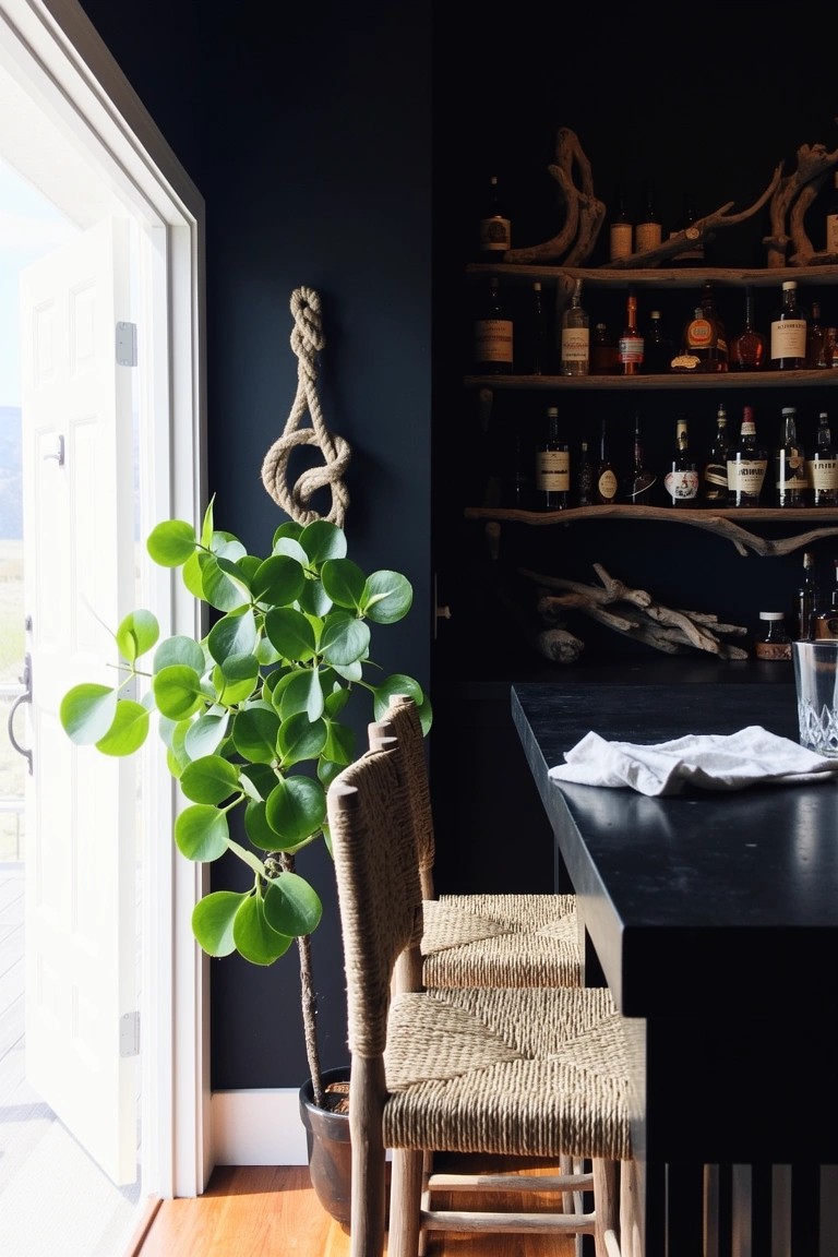 Black-walled bar area with knotted rope and potted plant on wall, driftwood shelves holding whiskey bottles, rattan stools at wooden bar counter, large windows open to outside