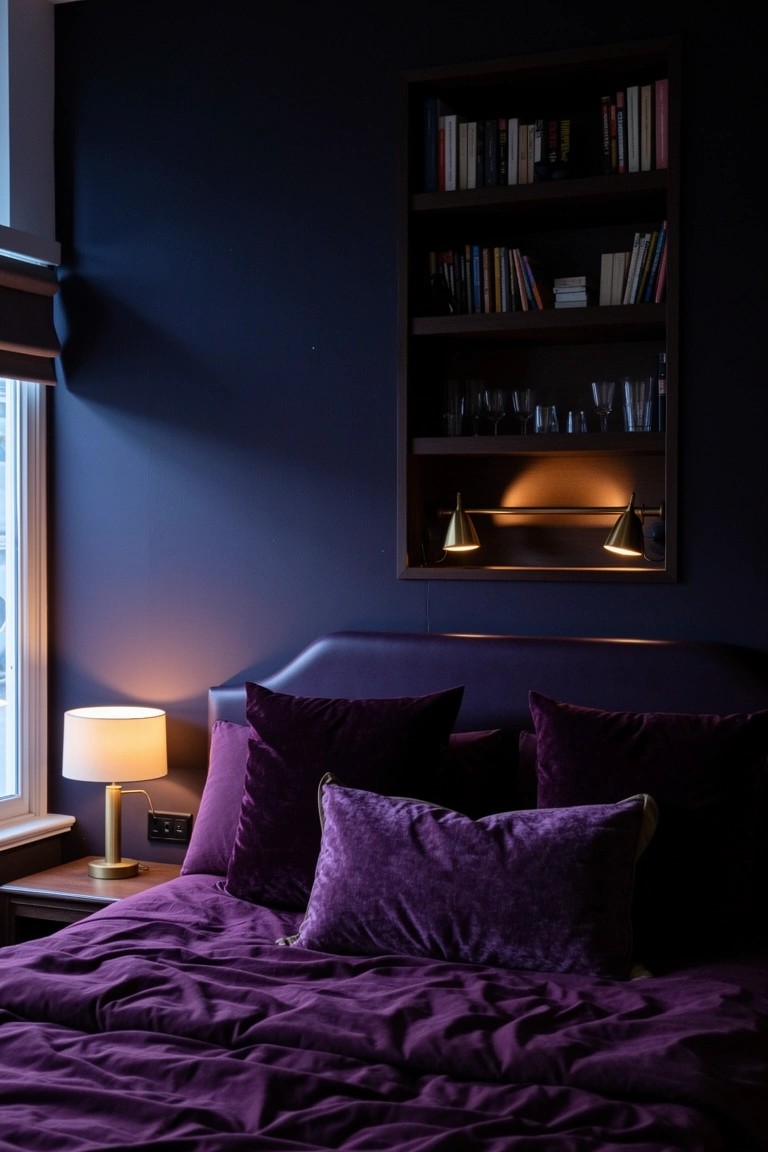 Navy bedroom wall with built-in shelves holding books and glassware above purple velvet bed and brass lights