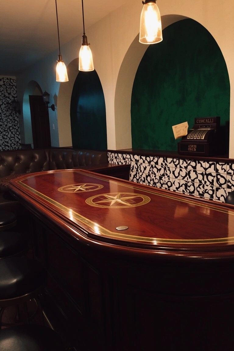 Home speakeasy bar in arched green alcove with wooden top, leather stools, and pendant lights
