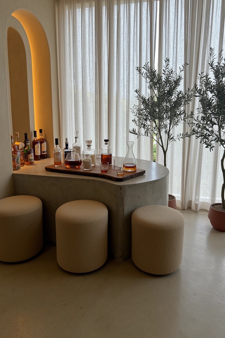 Round concrete table with whiskey bottles and glasses surrounded by three beige pouf stools in a neutral room with sheer curtains and potted olive trees