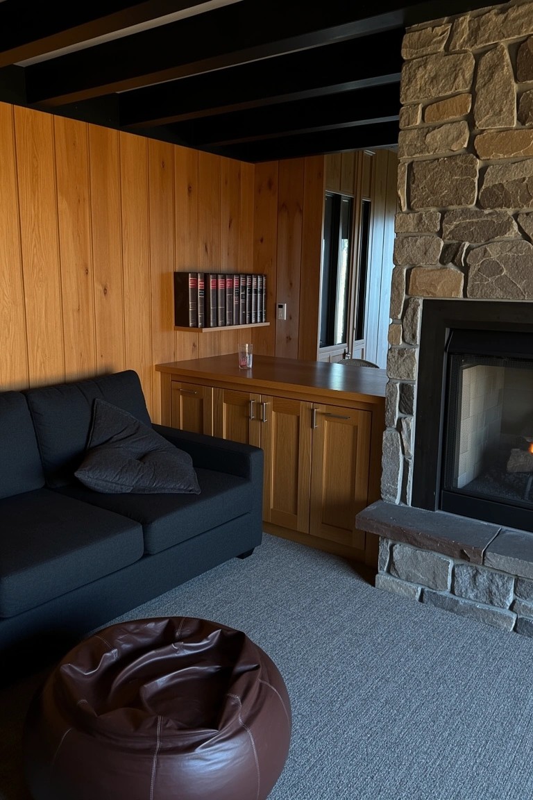 Man cave interior with warm wood panel walls, stone fireplace, black sofa, and built-in wood bar cabinet
