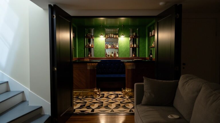 21 Stunning Speakeasy Room Ideas You Will Want To Try Compact speakeasy bar nook with green walls, brass-trimmed wooden counter, blue velvet bench, bottle shelves, and arched doorway from stairs