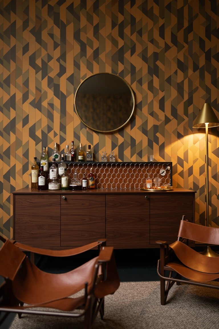 Orange and black geometric wallpaper behind a wooden bar cabinet with liquor bottles, round mirror, gold lamp, and leather sling chairs in a speakeasy nook