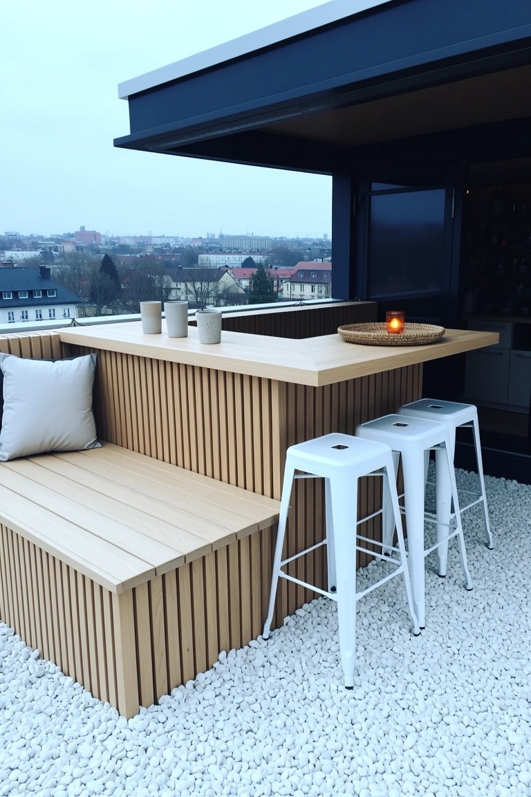 Light slatted wood bar counter and bench on rooftop terrace with white stools, cups, and candle, overlooking city buildings