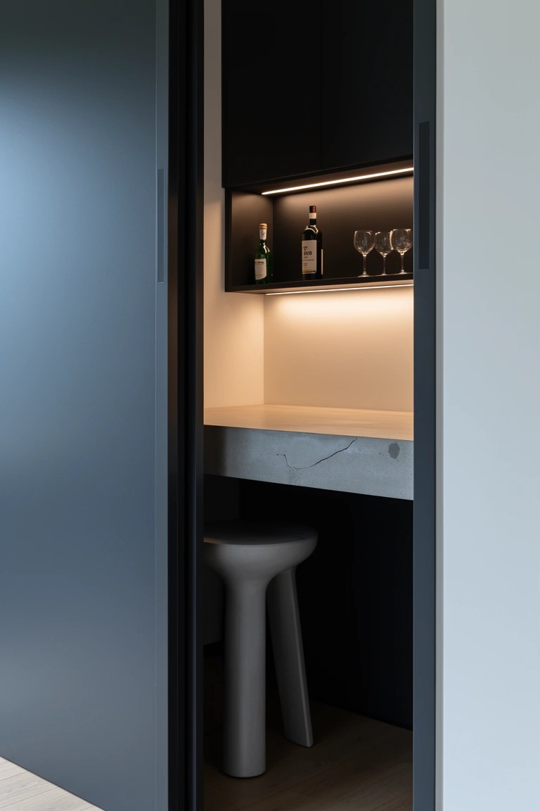 Recessed dark cabinet bar nook with backlit shelves holding whiskey bottles and glasses above a white marble counter and stool