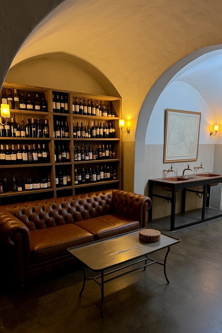 Leather tufted sofa in front of wooden wine racks lining arched beige walls, with wall sconces and a low table in a dimly lit lounge