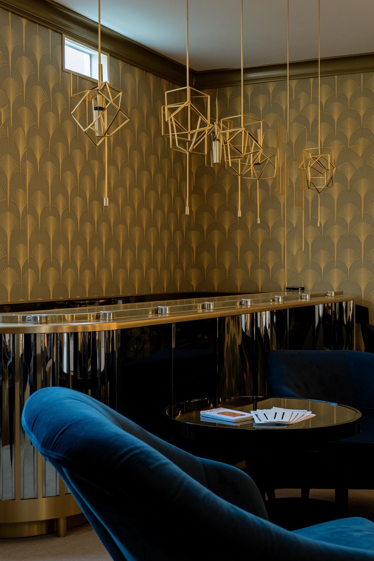Gold geometric pendant lights hanging over a shiny brass bar counter in a lounge with blue velvet chairs and patterned walls