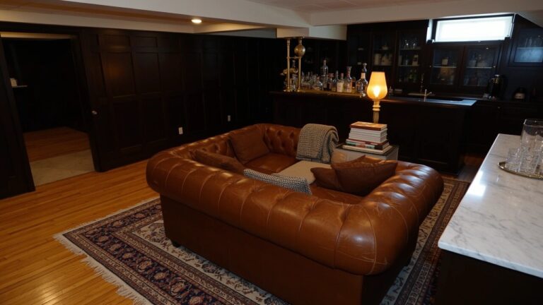 Dark wood paneled whiskey lounge with marble-top bar stocked with decanters and a tufted tan leather Chesterfield sofa with throw blanket