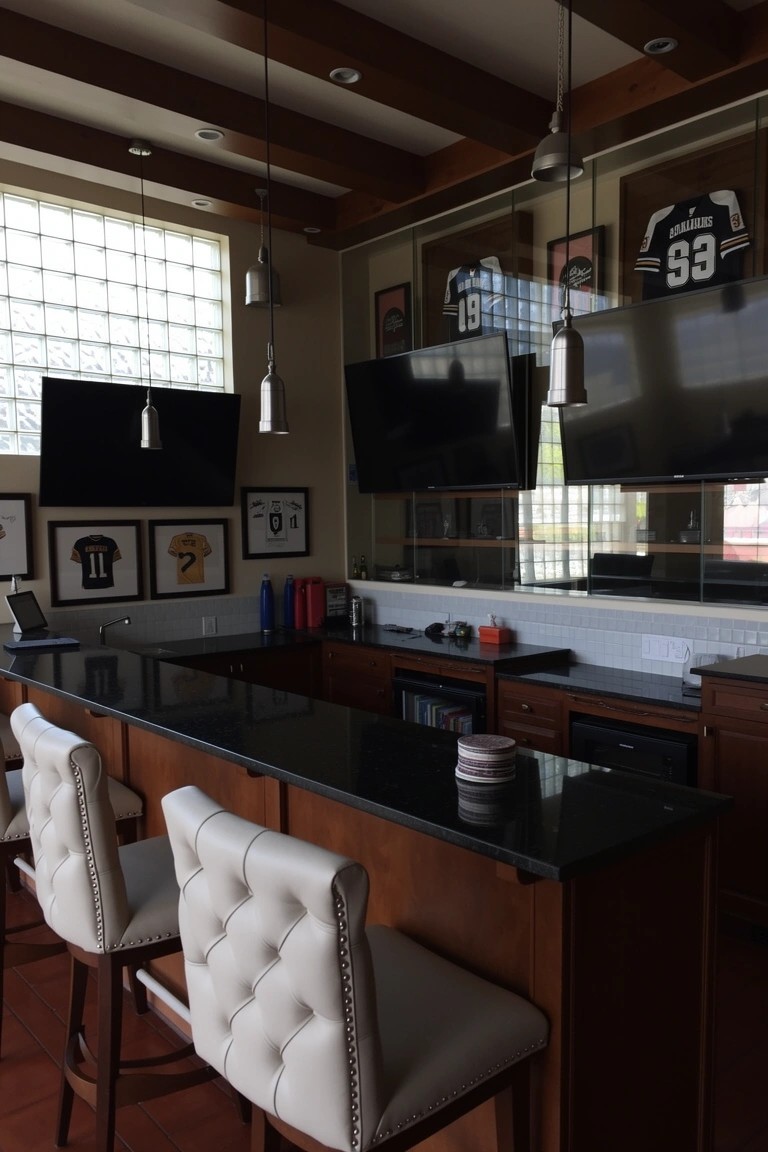 Man cave bar with dark wood cabinets, black granite countertop, white barstools, and sports jerseys on wood-paneled walls