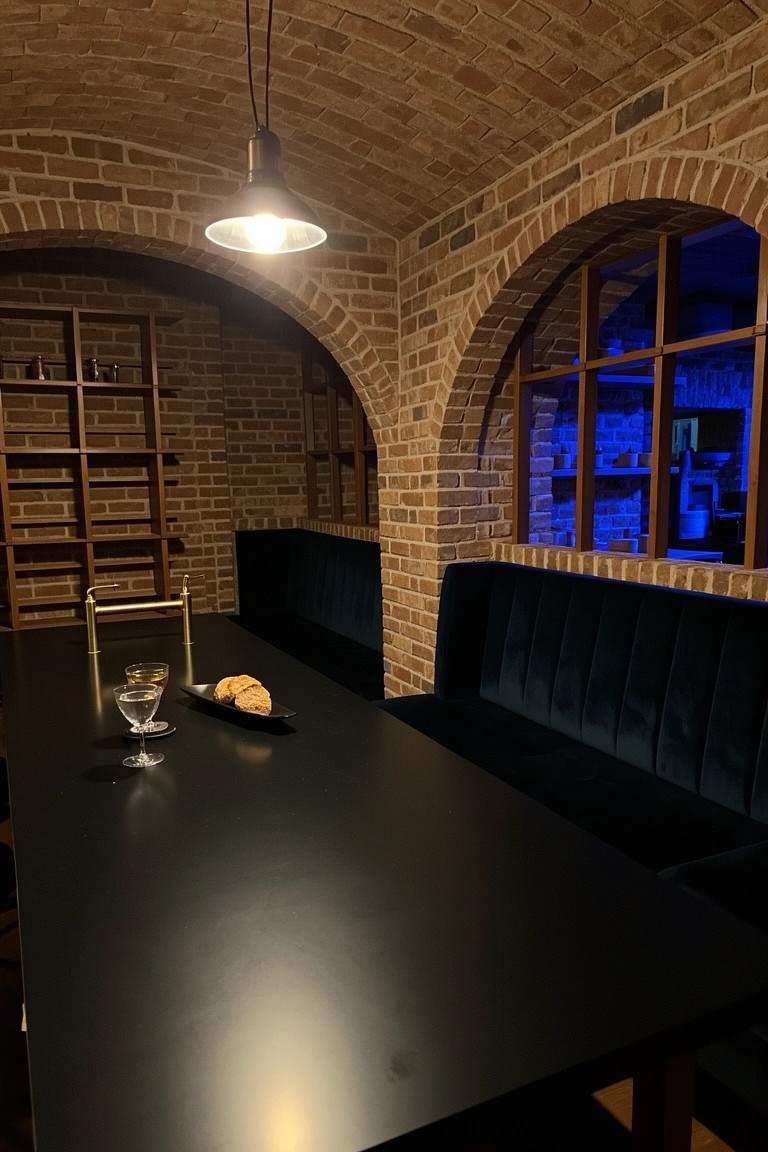 Brick-arched wine cellar with velvet banquette, black table, pendant light, and built-in wine racks