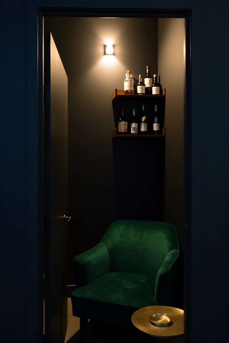 Narrow dark closet as whiskey nook with green velvet armchair, small round table, and wall shelves stocked with bottles