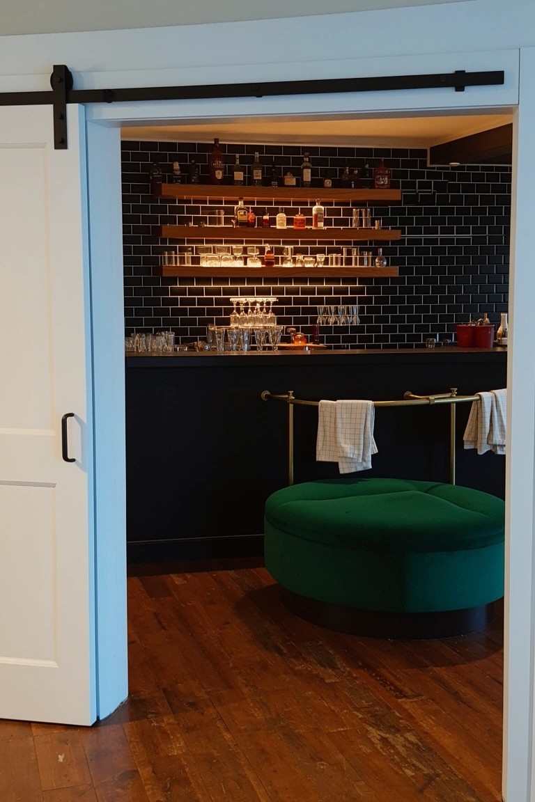 Home bar nook with backlit whiskey bottle shelves on black subway tile wall and green velvet pouf stool