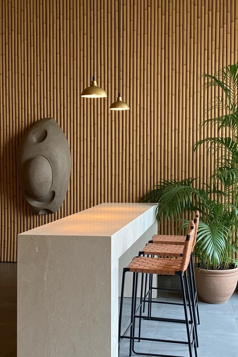 Sleek modern bar counter with bamboo wall paneling, white stone countertop, rattan stools, gold pendant lights, and potted palms