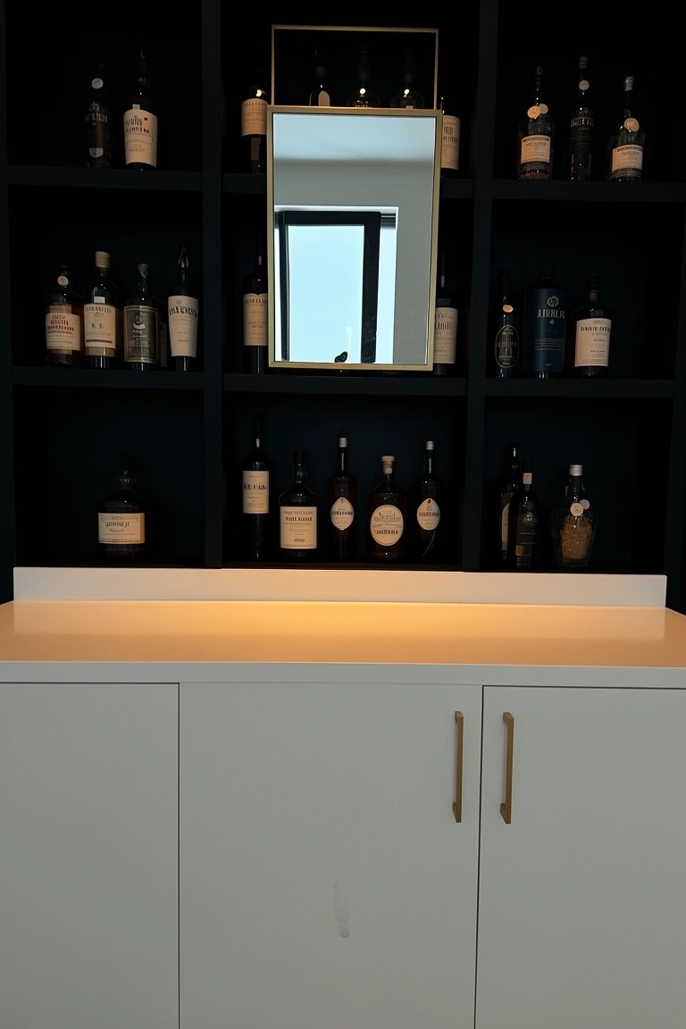 Dark open shelves lined with whiskey bottles and a central gold-framed mirror above white base cabinets in a modern bar area