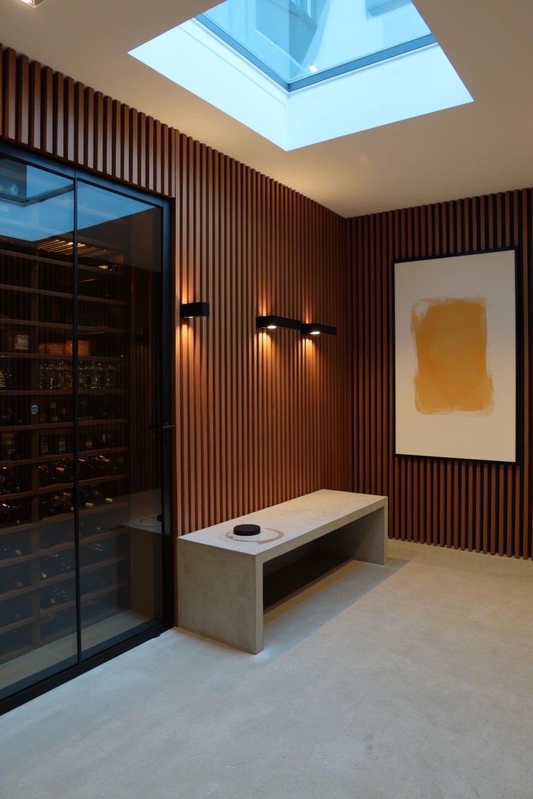 Modern whiskey room corner with vertical brown wood slat wall, glass-fronted wine storage, concrete bench, and wall lights