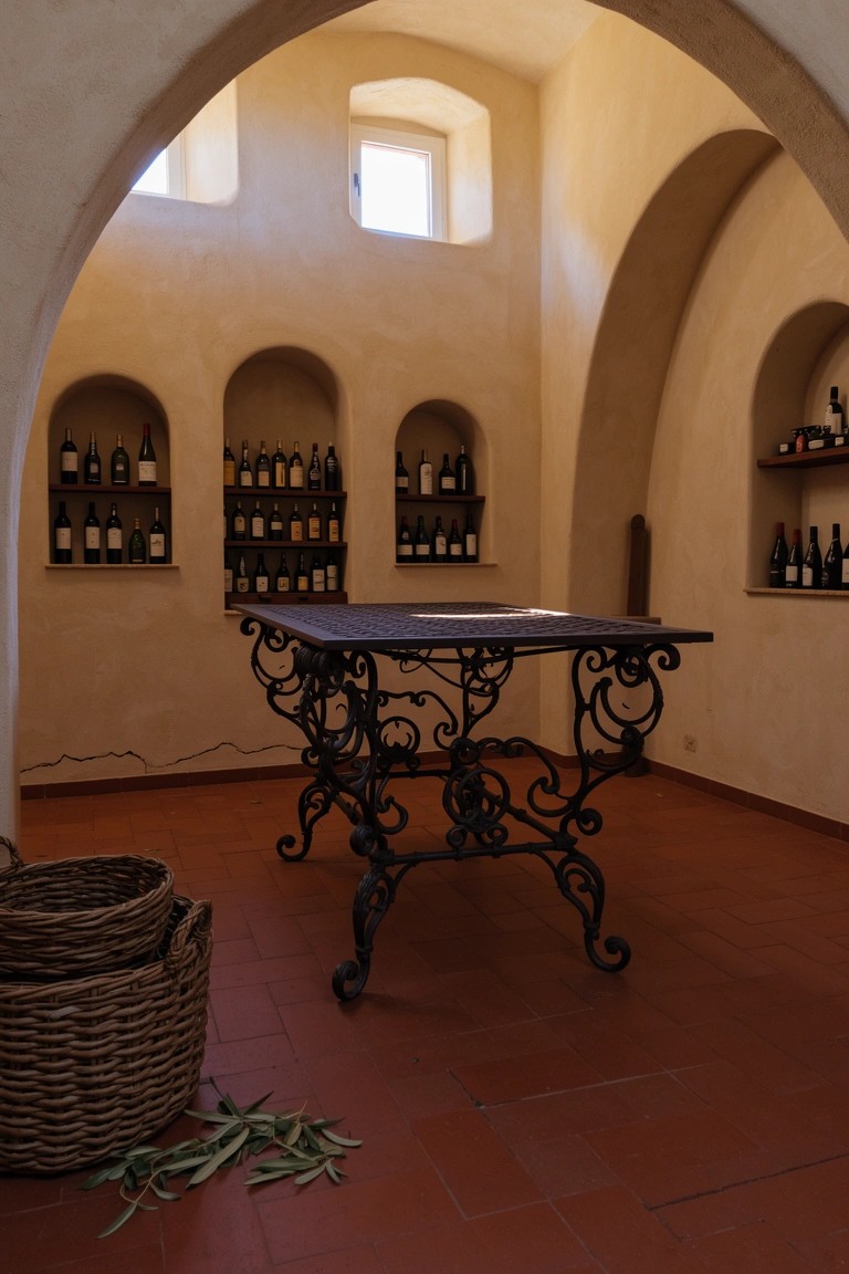 Rustic room with arched plaster walls holding rows of wine bottles around a central wrought iron table on terracotta tile floor