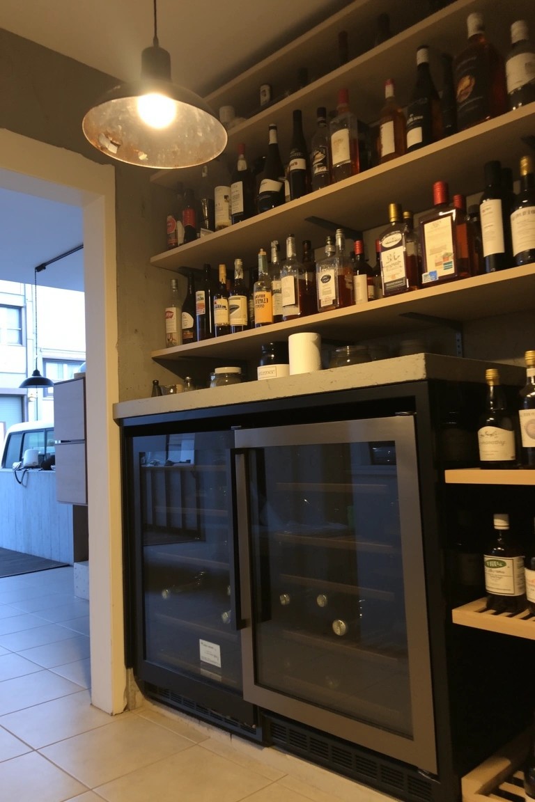 Wooden shelves filled with whiskey bottles above a black double-door beverage fridge in a home bar nook