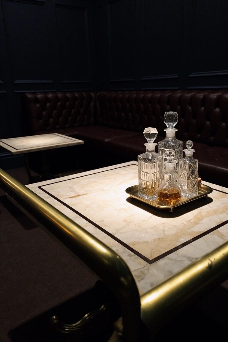 Brass bar cart with crystal decanters on marble table next to tufted leather booth in dark paneled basement room