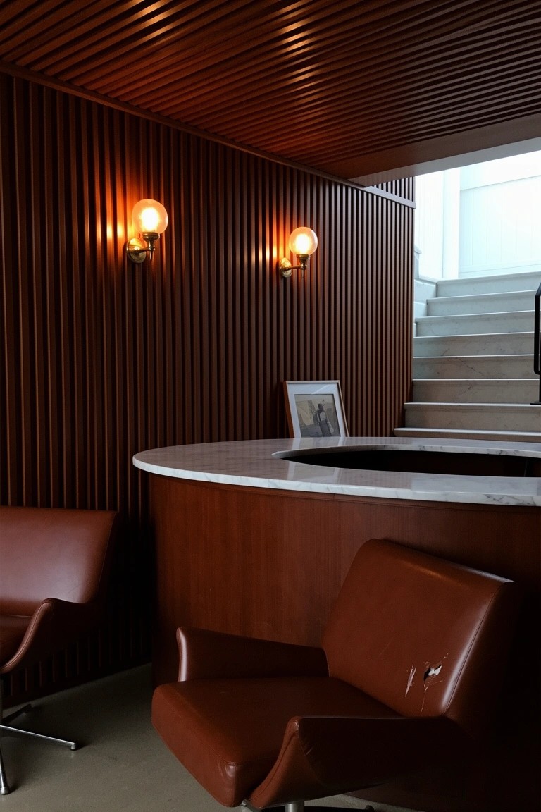 Curved white-topped wooden bar counter in a basement speakeasy with wood-paneled walls, leather swivel chairs, and warm sconce lighting