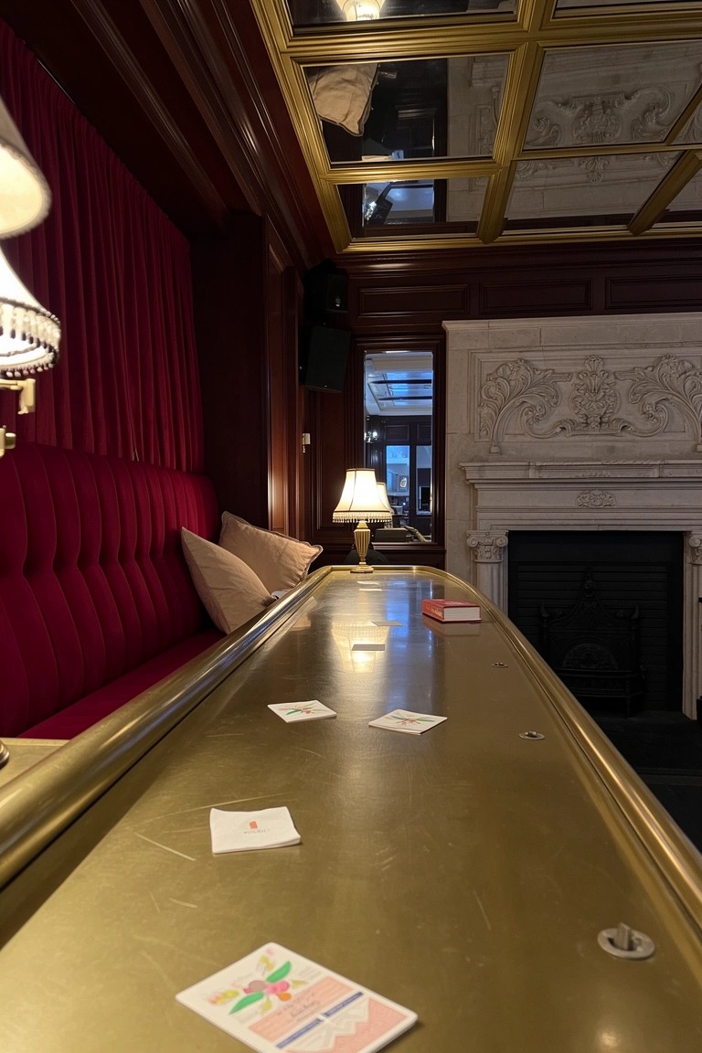 Gold bar counter with scattered cards in a wood-paneled basement speakeasy lounge featuring red velvet seating and a fireplace